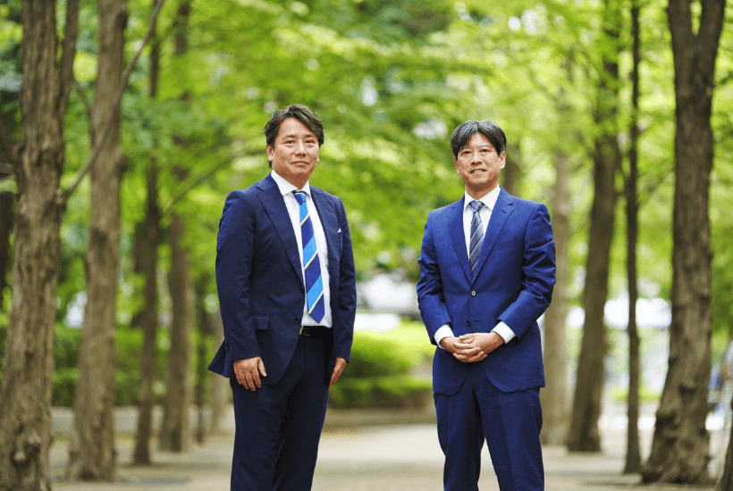 Two men in suits standing outdoors on a tree-lined path.